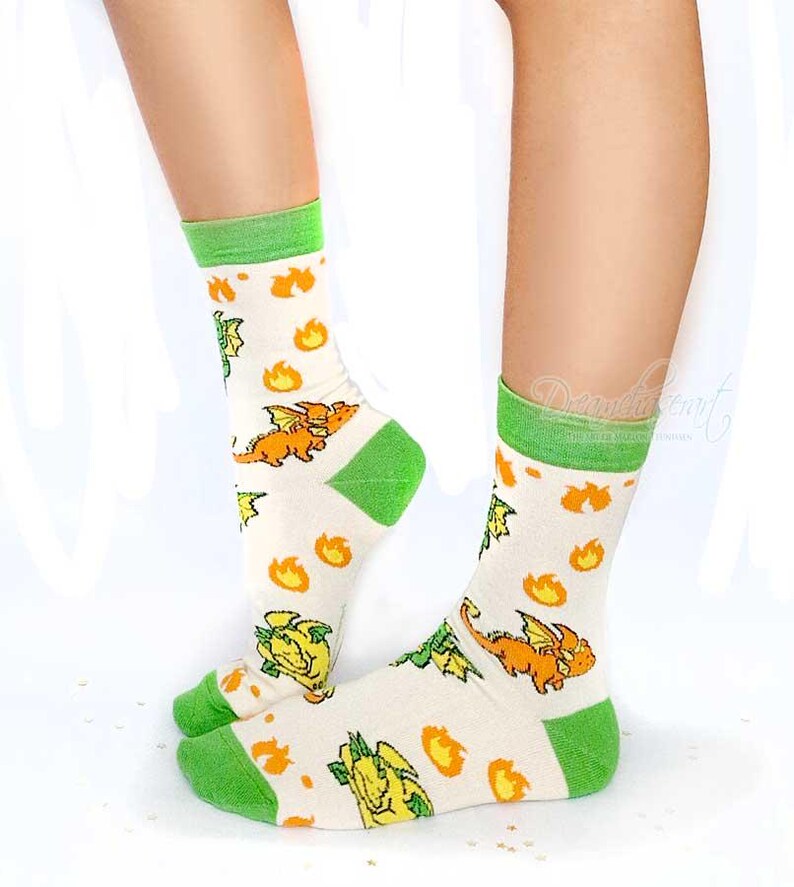 May include: Cream socks with green cuffs, heels, and toes. The socks display a repeating pattern of orange dragons and yellow flames. The socks are knee-high and made of a soft material.