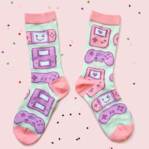 Pode incluir: Um par de meias rosa e verde com um padrão de consoles e controladores de videogame roxos. As meias têm uma bainha rosa e um fundo verde.