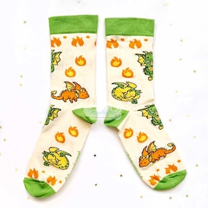 May include: A pair of cream socks with green cuffs and toes. The socks display a repeating pattern of orange and yellow cartoon dragons and flames. These socks are designed for comfort and style.