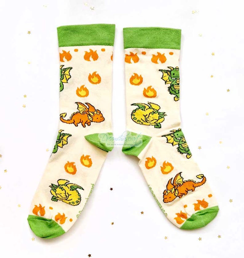 May include: A pair of cream socks with green cuffs and toes. The socks display a repeating pattern of orange and yellow cartoon dragons and flames. These socks are designed for comfort and style.