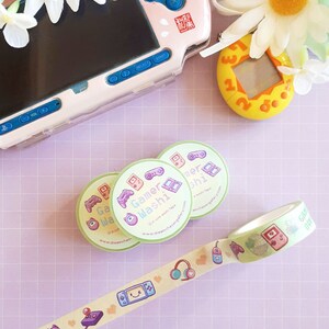 May include: A light green washi tape roll and three matching stickers with gaming-themed illustrations. The tape features game controllers, headphones, and other icons. A pink and blue handheld gaming device and a yellow digital pet toy are also visible.