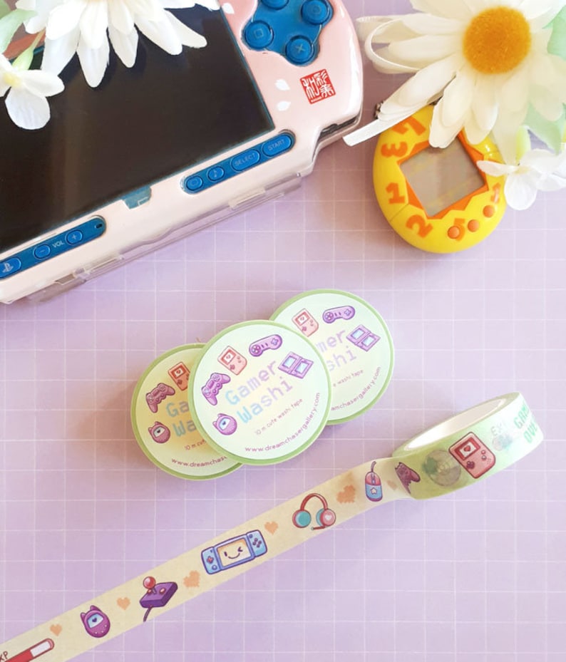 May include: A light green washi tape roll and three matching stickers with gaming-themed illustrations. The tape features game controllers, headphones, and other icons. A pink and blue handheld gaming device and a yellow digital pet toy are also visible.