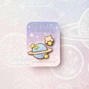 Peut inclure: Épinglette en émail Pastel Galaxy représentant une planète bleue et rose avec un anneau, entourée d'étoiles jaunes. L'épingle est sur une carte blanche avec le texte "Pastel Galaxy" et une adresse de site web.