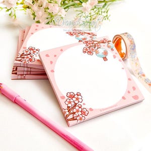 May include: A stack of square pink sticky notes featuring a large white circular writing area, decorated with illustrated cherry blossom branches and hanging lanterns. A pink pen and a roll of floral patterned washi tape are placed nearby.