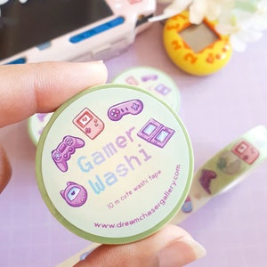 May include: A roll of Gamer Washi tape with a light green background, featuring game-themed illustrations in purple and pink. The tape includes text that reads "Gamer Washi" and "10 m cute washi tape".