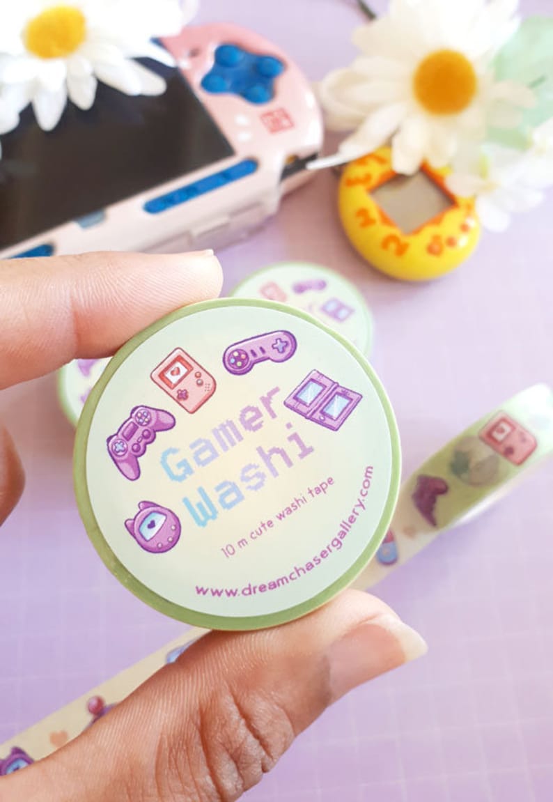 May include: A roll of Gamer Washi tape with a light green background, featuring game-themed illustrations in purple and pink. The tape includes text that reads "Gamer Washi" and "10 m cute washi tape".