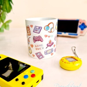 Pu&ograve; includere: Una tazza in ceramica bianca con un design colorato di icone di videogiochi retr&ograve;, tra cui controller, cuffie e una cartuccia di gioco. Il testo "GAME OVER" &egrave; stampato in lettere pixelate sulla tazza.