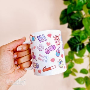 Pu&ograve; includere: Tazza in ceramica bianca con un design rosa, blu e verde con varie icone di videogiochi retr&ograve;, tra cui un joystick, una cartuccia di gioco, un floppy disk, un cuore e il testo "GAME OVER".