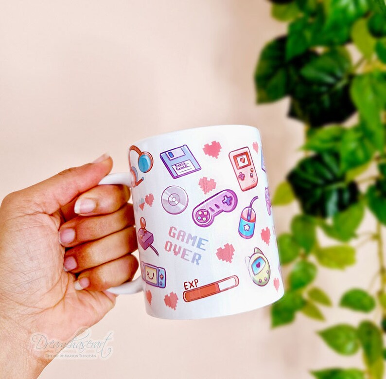 Pu&ograve; includere: Tazza in ceramica bianca con un design rosa, blu e verde con varie icone di videogiochi retr&ograve;, tra cui un joystick, una cartuccia di gioco, un floppy disk, un cuore e il testo "GAME OVER".