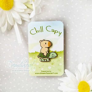 Puede incluir: Un pin de esmalte dorado con un capibara de dibujos animados montando una tortuga. El pin está en una tarjeta blanca con el texto "Chill Capy" y "Dreamchaserart" en letras verdes y negras.