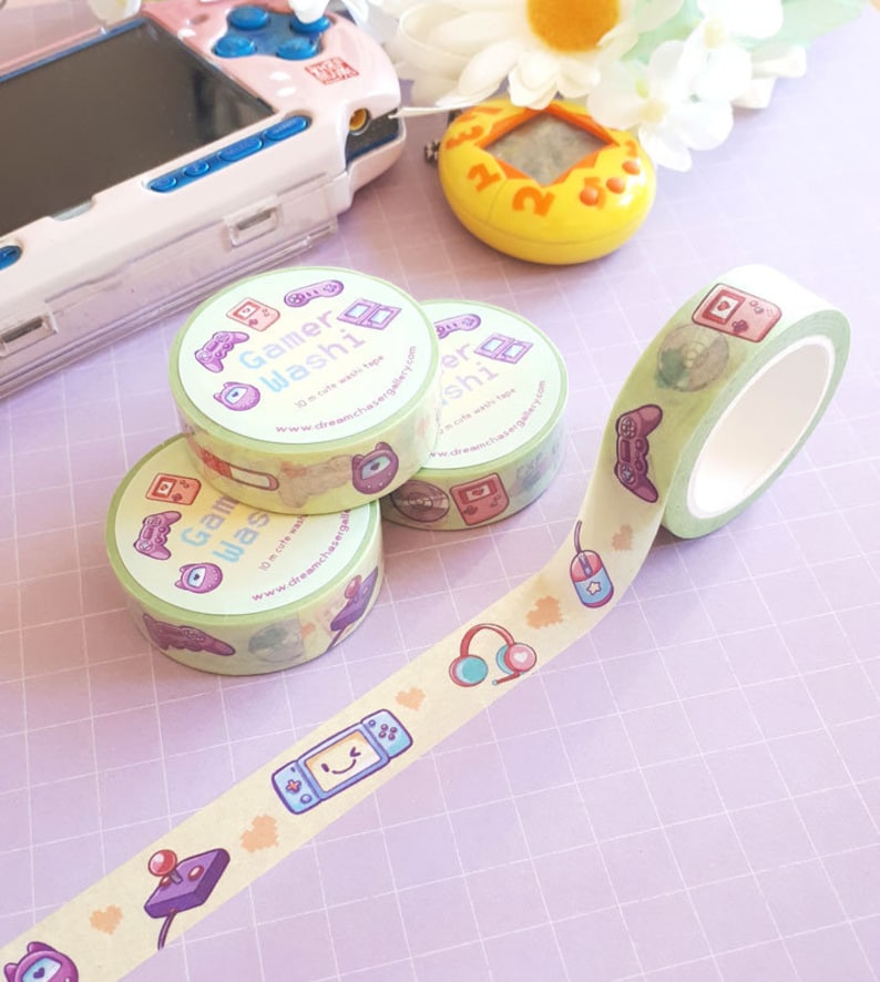 May include: A collection of Gamer Washi tape rolls with a light green background and various gaming-themed illustrations. The tape features images of game controllers, headphones, and other gaming icons. The text "Gamer Washi" is printed on the tape rolls.