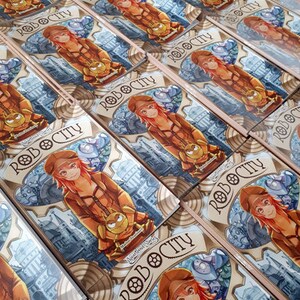 May include: Several copies of a card game box with a steampunk illustration. The artwork shows a red-haired character in a brown outfit, holding a small robot. The game title "ROBOCITY" is displayed in a banner.