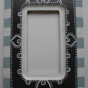 May include: A black and white light switch cover with a decorative design. The cover has a blue and white striped border.