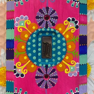 Puede incluir: Una cubierta de interruptor de luz pintada a mano con un diseño vibrante y colorido. La cubierta presenta un fondo rosa brillante con patrones florales turquesa, morado, naranja y amarillo. La placa del interruptor es de tamaño estándar.