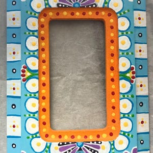 May include: Hand-painted rectangular picture frame with a vibrant floral design. The frame features a light blue background with white, purple, and yellow floral accents. The inner frame is orange with red dots. Decorative and colorful.