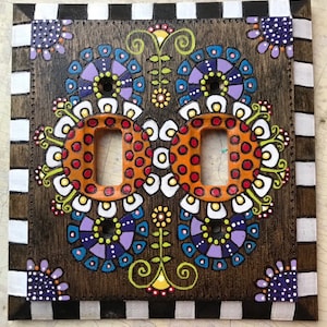 May include: Hand-painted wooden double light switch cover with a floral design. The cover features orange, purple, and white floral patterns, with a dark brown background. The edges are decorated with a black and white checkered pattern.