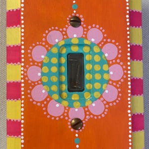 以下が含まれることがあります： カラフルなマンダラ模様が手描きされたライトスイッチカバー。カバーはオレンジ色で、中央にはターコイズブルー、イエロー、ピンクのマンダラが描かれています。スイッチは白い枠とピンクとイエローのストライプの枠で囲まれています。