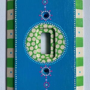 May include: A rectangular light switch cover plate featuring a bright blue center with hand-painted dot art patterns in pink, light blue, and white. The border has alternating green and cream stripes with small blue dots on each cream section.