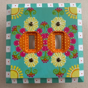 May include: A square double light switch wall plate featuring a hand-painted teal background with a colorful floral pattern in yellow, orange, and pink. The border has white and teal stripes with orange dots, and two metal screws are visible.