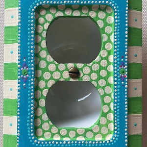 May include: A hand-painted light switch cover with a green and white polka dot pattern, a turquoise blue frame, and white and green stripes on the outer edge.