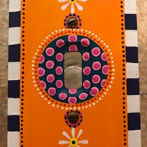 May include: A rectangular light switch plate featuring a bright orange center with hand-painted white daisy motifs, a dark blue circular accent with pink dots, and a border of navy blue and white stripes along the outer edges with two metal screws.