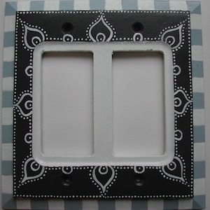 May include: A decorative double light switch cover. The cover has a black border with white dotted and teardrop-shaped designs. The border is framed by a light blue and white checkered pattern.