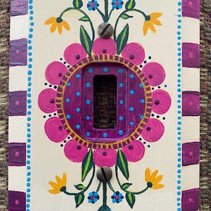 Pode incluir: Tampa de interruptor de luz pintada à mão com um design floral. A tampa apresenta um fundo branco com uma borda listrada roxa e branca. O centro tem um círculo roxo com flores rosa e pontos azuis. Flores amarelas e azuis também estão presentes.