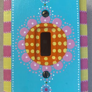 May include: A hand-painted light switch cover with a vibrant design. The central panel is turquoise, adorned with an orange circle featuring yellow polka dots. Pink and white dot patterns and a pink and yellow checkered border complete the design.