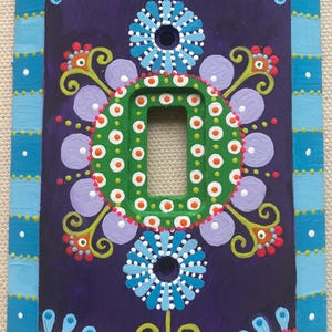 May include: A rectangular light switch cover plate featuring a dark purple background with a hand-painted design. The border has light blue and dark blue stripes with white dots. The center displays green, light blue, and lavender floral motifs.