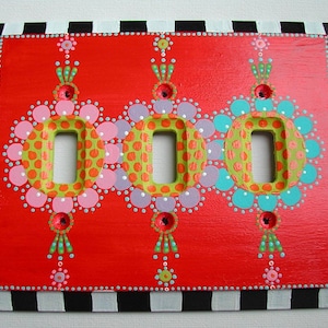 May include: A hand-painted light switch cover with three round designs in shades of pink, green, blue, and purple. The designs are surrounded by black and white polka dots and a black and white checkered border.