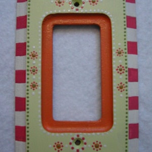 May include: Decorative light switch cover with a rectangular opening. The cover features a light green center with an orange inner frame. The outer border is white with red and pink stripes. The cover is decorated with small dots and floral accents.