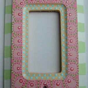 May include: A pink light switch cover with a decorative border of white, green, and blue dots. The border features a repeating pattern of pink circles with white dots.