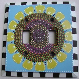 Puede incluir: Una tapa de interruptor doble de color azul claro con un borde a cuadros blanco y negro. El centro de la tapa presenta un diseño de flor amarilla con un centro de lunares blanco y negro.