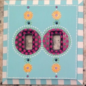 May include: A decorative light switch cover with a light blue background. The cover features two rectangular switch openings surrounded by pink polka-dotted circles. Yellow and purple flower designs and white dotted borders add to the whimsical design.