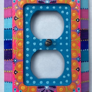 May include: A rectangular electrical outlet wall plate featuring a hand-painted design with a bright blue center dotted with white, surrounded by a vibrant pink border with orange, purple, and blue floral-inspired patterns and a metal screw.