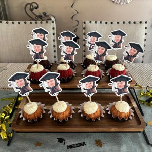 May include: A two-tiered cake stand with cupcakes decorated with graduation caps and personalized photos. The cupcakes are topped with chocolate and vanilla frosting. The stand is made of wood and has a black metal frame.