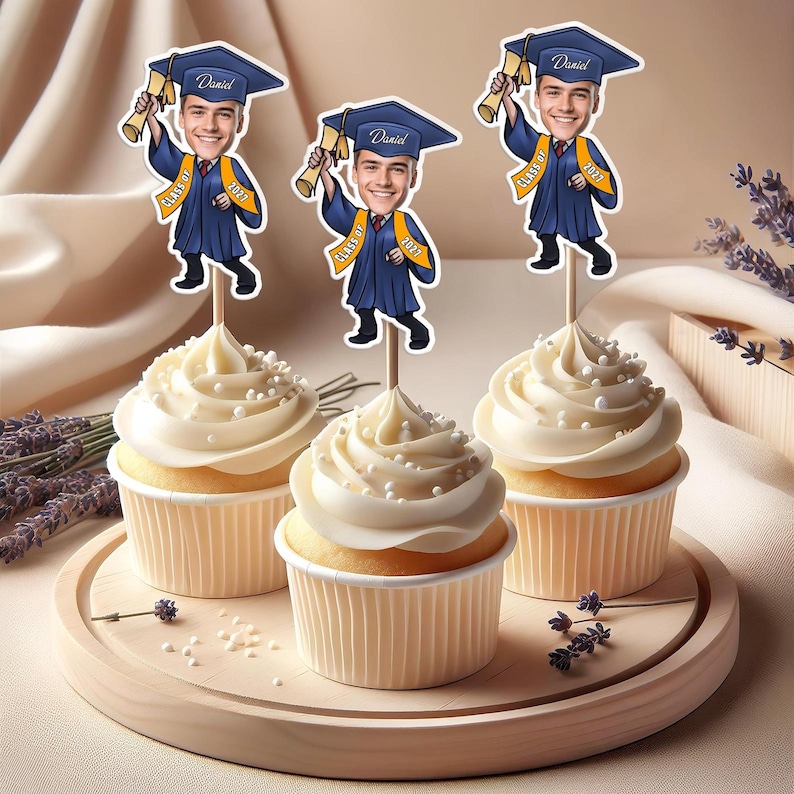 May include: Three cupcakes with white frosting and white sprinkles topped with graduation themed cake toppers featuring a cartoon graduate wearing a cap and gown, holding a diploma, and the text "Class of 2023" and "Daniel".