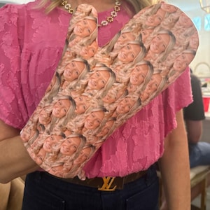 May include: A white oven mitt with a repeated pattern of a woman's face. The mitt is being held by a person wearing a pink shirt and blue jeans.