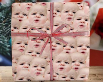 May include: A wrapped gift box featuring a repeating pattern of baby faces. The wrapping paper is tied with a red ribbon and bow. The gift box is on a wooden surface.