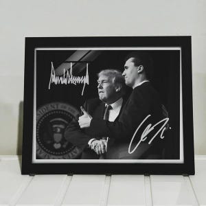 May include: Framed black and white photograph featuring two men, one giving a thumbs up. The photograph is signed with white ink. The presidential seal is visible in the background.