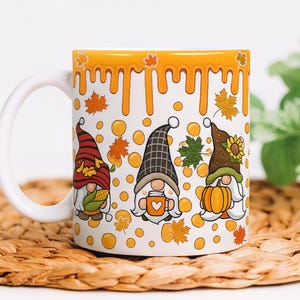 May include: White ceramic mug featuring three whimsical gnomes in autumn attire. The gnomes are adorned with hats, holding a pumpkin, a mug, and corn. The mug has a yellow dripping design at the top and is decorated with fall leaves.