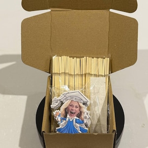 May include: A brown cardboard box with a black base is open and contains a graduation cake topper with a woman's face on it. The topper says "Lint Grace" and "Class of 2023". The box also contains a package of wooden sticks and a package of adhesive dots.