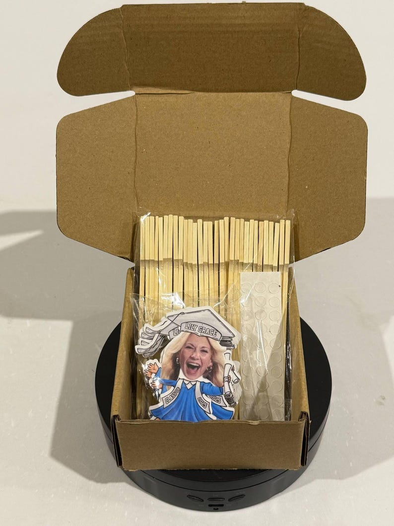 May include: A brown cardboard box with a black base is open and contains a graduation cake topper with a woman's face on it. The topper says "Lint Grace" and "Class of 2023". The box also contains a package of wooden sticks and a package of adhesive dots.