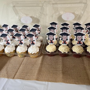May include: Two clear plastic containers filled with cupcakes decorated with graduation caps and faces. The cupcakes are topped with white frosting and are arranged in rows.