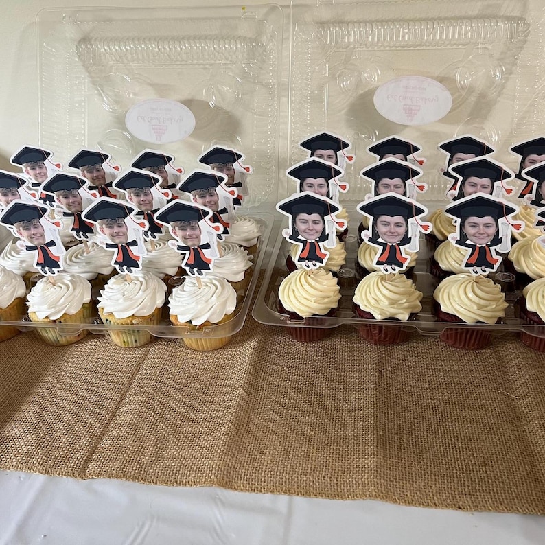 May include: Two clear plastic containers filled with cupcakes decorated with graduation caps and faces. The cupcakes are topped with white frosting and are arranged in rows.