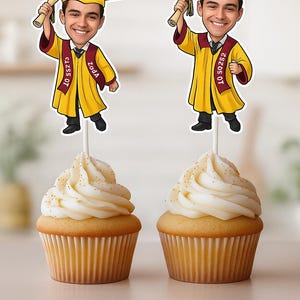 May include: Two yellow graduation cupcakes with a cartoon graduate topper. The topper is a caricature of a person wearing a graduation cap and gown, holding a diploma. The text on the topper says "Daniel" and "Class of 2023".