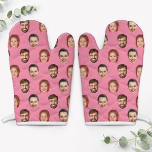 May include: A pair of pink oven mitts with a quilted pattern. The mitts are covered in a repeating pattern of smiling faces. The mitts have a white loop for hanging and are designed for kitchen use.