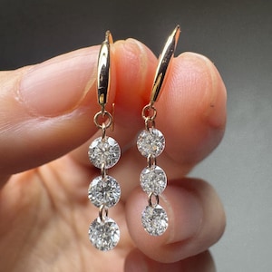 May include: A pair of gold drop earrings, each with three clear, sparkling round gemstones. The earrings hang from a gold hook. The gemstones are of varying sizes, creating a cascading effect.