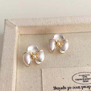 Pode incluir: Um par de brincos de tarracha em forma de flores de tr&ecirc;s p&eacute;talas. Cada brinco apresenta p&eacute;talas brancas peroladas com um centro dourado e uma pequena conta branca redonda no meio, criando um design floral delicado e sim&eacute;trico.
