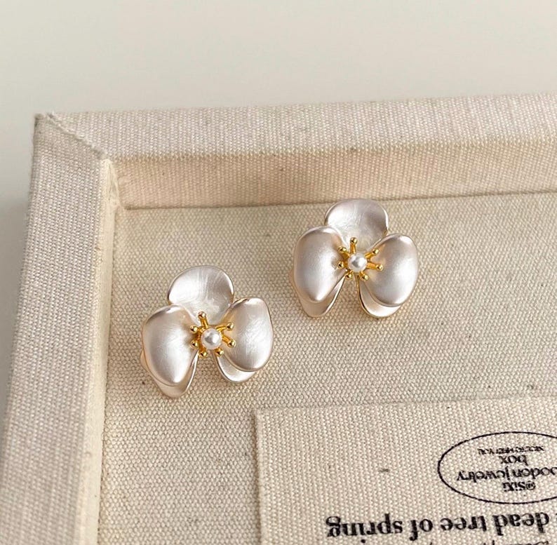 Pode incluir: Um par de brincos de tarracha em forma de flores de tr&ecirc;s p&eacute;talas. Cada brinco apresenta p&eacute;talas brancas peroladas com um centro dourado e uma pequena conta branca redonda no meio, criando um design floral delicado e sim&eacute;trico.